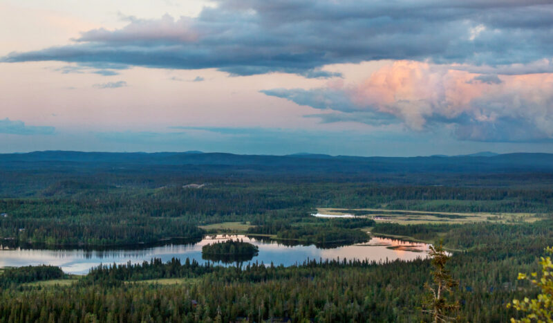 Mäeltä kuvattu maisema, jossa metsiä, järviä ja pilviä ilta-auringossa.