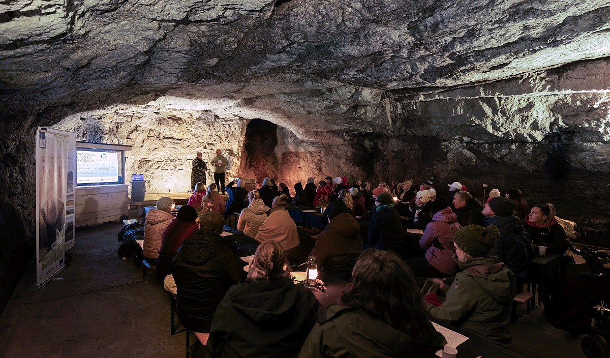 Hämärä kiviseinäinen luola, jossa etualaa kuulijoita istumassa pöytien äärellä ja taustalla koroke, jossa seisoo kaksi puhujaa.