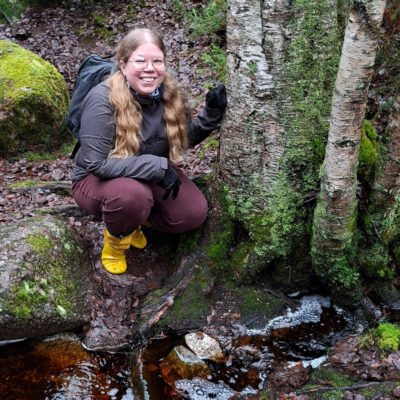Sara Käkelä kyykistyneenä pienen metsäpuron takana, ympärillä koivuja ja sammaleisia kiviä .