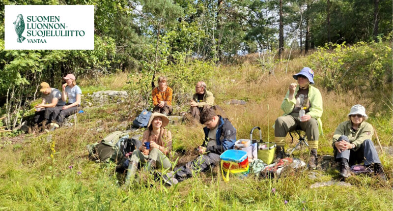 Ihmisiä istumassa niityn reunan kalliolla, taustalla metsää.